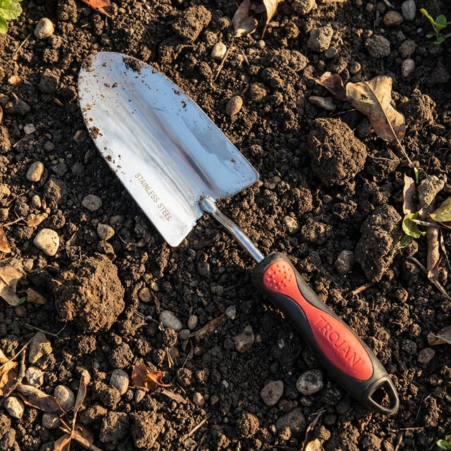 The Trojan Stainless Steel Hand Trowel, featuring a red and black handle, is perfect for potting and planting. It is shown resting on soil with small rocks and leaves.
