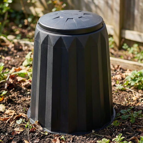 A Tumbleweed 150L Gedye Compost Bin sits on garden soil, surrounded by dry leaves and a wooden fence—this compact compost bin is perfect for small spaces.