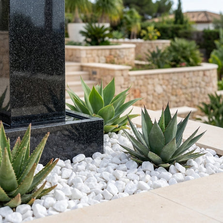Agave plants are bordered by Ki Carma 20kg 10 to 20mm Drainage Gravel and white pebbles beside a black stone structure in a beautifully landscaped garden.