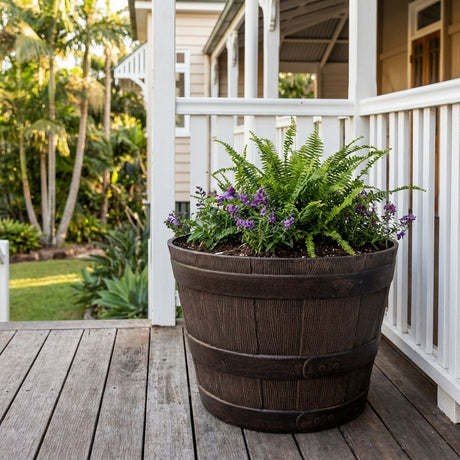 The Walnut Whiskey Barrel, available in various sizes, serves as a rustic outdoor planter filled with ferns and purple flowers, adding vibrant charm to any porch surrounded by greenery.