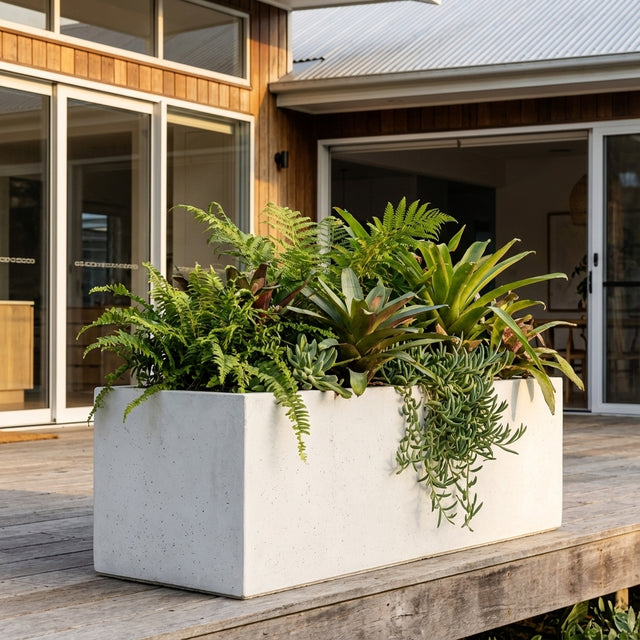 A White Emmerson Trough planter, available in various sizes, filled with green plants sits on a wooden deck outside a contemporary house.