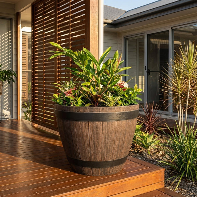 The Wine Barrel Planter - 800mm, filled with lush green plants, sits on a wooden deck beside a modern house with sliding doors.