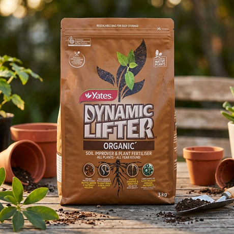 A 3kg bag of Yates Dynamic Lifter Soil Improver and Plant Fertiliser sits on a wooden table next to potted plants and soil.